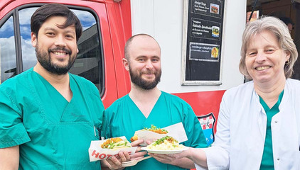Zwei Pflegekräfte in blauer Funktionskleidung eine Ärztin in weißer Funktionskleidung stehen mit Essen in den Händen vor einem Food-Truck am Internationalen Tag der Pflegenden im Immanuel Klinikum Bernau Herzzentrum Brandenburg.