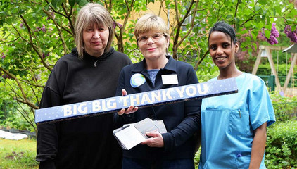 Zwei Pflegekräfte stehen mit Sabine Kalkhoff, Gesamtleiterin Residenz am Wiesenkamp und Gesellschaft für Diakonie, beim Internationalen Tag der Pflegenden mit einem großen Querschild mit der Aufschrift "Big Big Thank You" vor einem Fliederbaum auf dem Außengelände des Feierabendhauses Volksdorf in Hamburg.