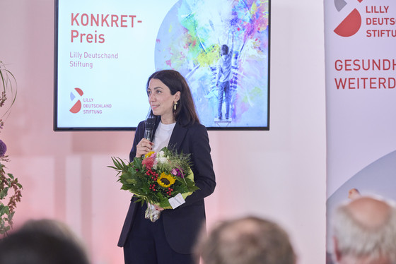 Dr. Viyan Sido steht mit Blumenstrauß auf der Bühne und spricht in ein Mikrofon Dr. Viyan Sido steht mit Blumenstrauß auf der Bühne und spricht in ein Mikrofon