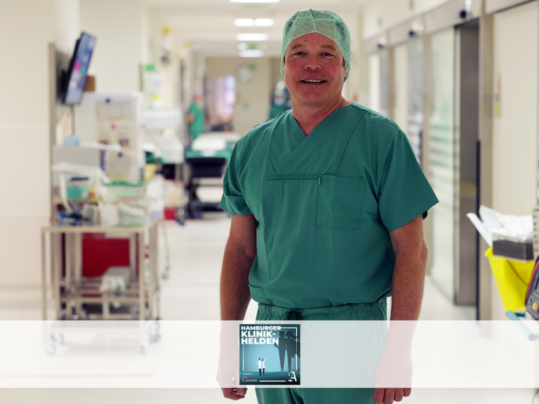 Dr. Ralf Bader, Leitender Oberarzt in der Klinik für Herzchirurgie im Albertinen Krankenhaus, blickt in grüner OP-Kleidung mit Kopfhaube in einem Krankenhausflur in die Kamera und war zu Gast im Podcast von Abendblatt und freigemeinnützigen Krankenhäusern "Hamburger Klinikhelden" zum Thema Bypass-Operationen.